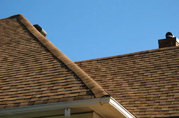 Well-maintained residential roof with brown asphalt shingles and two chimneys, highlighting quality installation and routine upkeep under a clear blue sky.