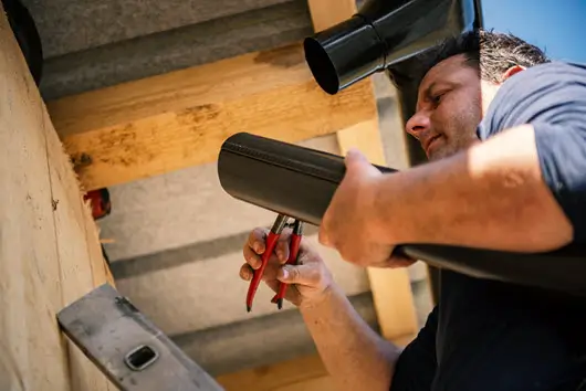 Man wearing a dark t-shirt, focused on repairing a black gutter with red-handled pliers. The scene shows a building's wooden eaves, a ladder, and a bright blue sky, illustrating hands-on gutter maintenance.