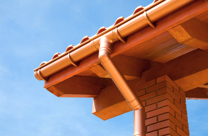 Low-angle view of a copper gutter system with a horizontal gutter and vertical downspout, set against a terracotta-tiled roof, wooden eaves, a brick pillar, and a clear blue sky.