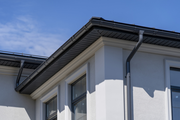 Close-up of a modern building's roofline with a dark gray rain gutter system, matching downspout, and decorative white facade, set against a clear blue sky with light clouds.