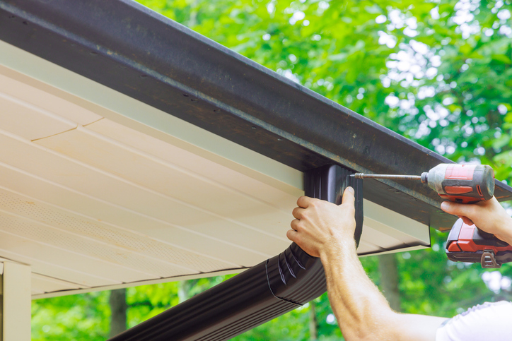 Close-up of a person installing a dark brown downspout onto a seamless gutter system, using a power drill, with a beige soffit and green foliage in the background.