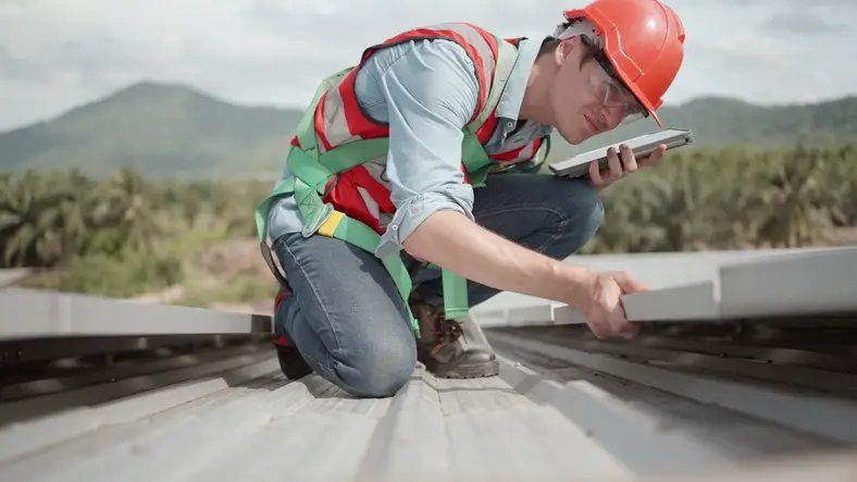 A Roofing inspector in safety gear, inspecting metal roof panels with a tablet in Logan, UT