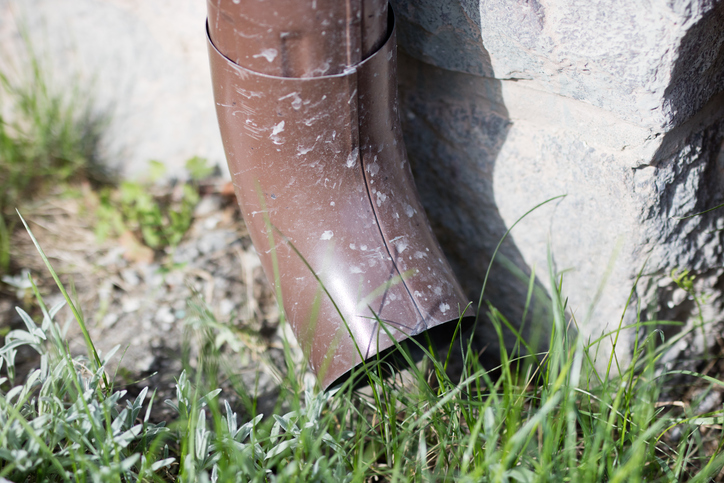 Close-up of a brown downspout with visible wear, curving towards the ground near a stone foundation, surrounded by grass and vegetation, emphasizing the importance of downspout unclogging.